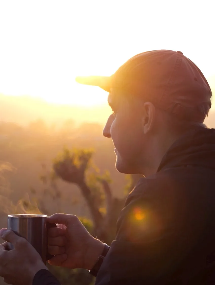 A man in a cap holds a steaming mug and watches the sun rise over wooded hills