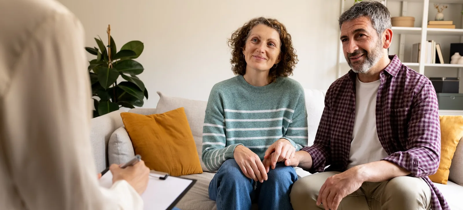 A couple sits close together on a sofa, smiling as they talk with a coach who is taking notes